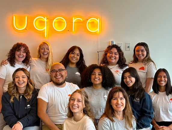 Group of people posing in front of a neon 'uqora' sign.