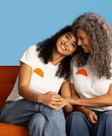 Two smiling women in white Uqora t-shirts sitting close together on an orange couch